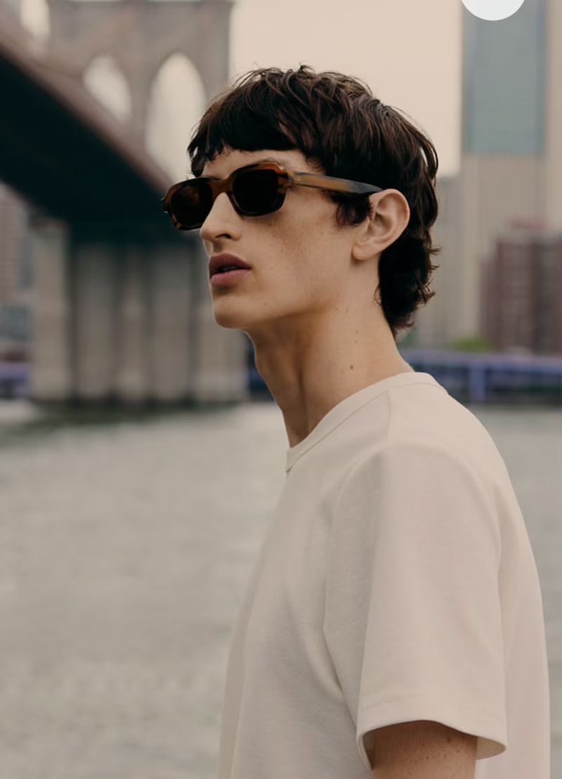 A young man wearing brown sunglasses and a white t-shirt stands near a body of water with a bridge in the background during the daytime.