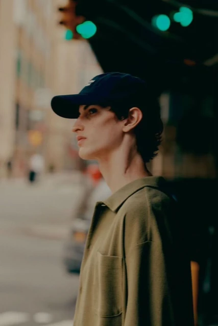 Young adult with a baseball cap and khaki shirt in a city street setting with buildings and streetlights. Medium shot, eye-level view.