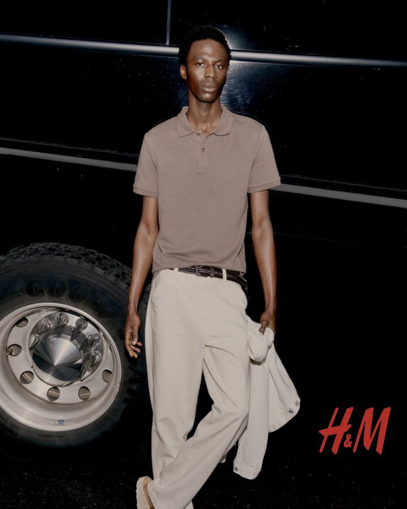 Adult male poses in front of a tour bus wearing a brown polo shirt and light tan dress pants for an H&M fashion advertisement.
