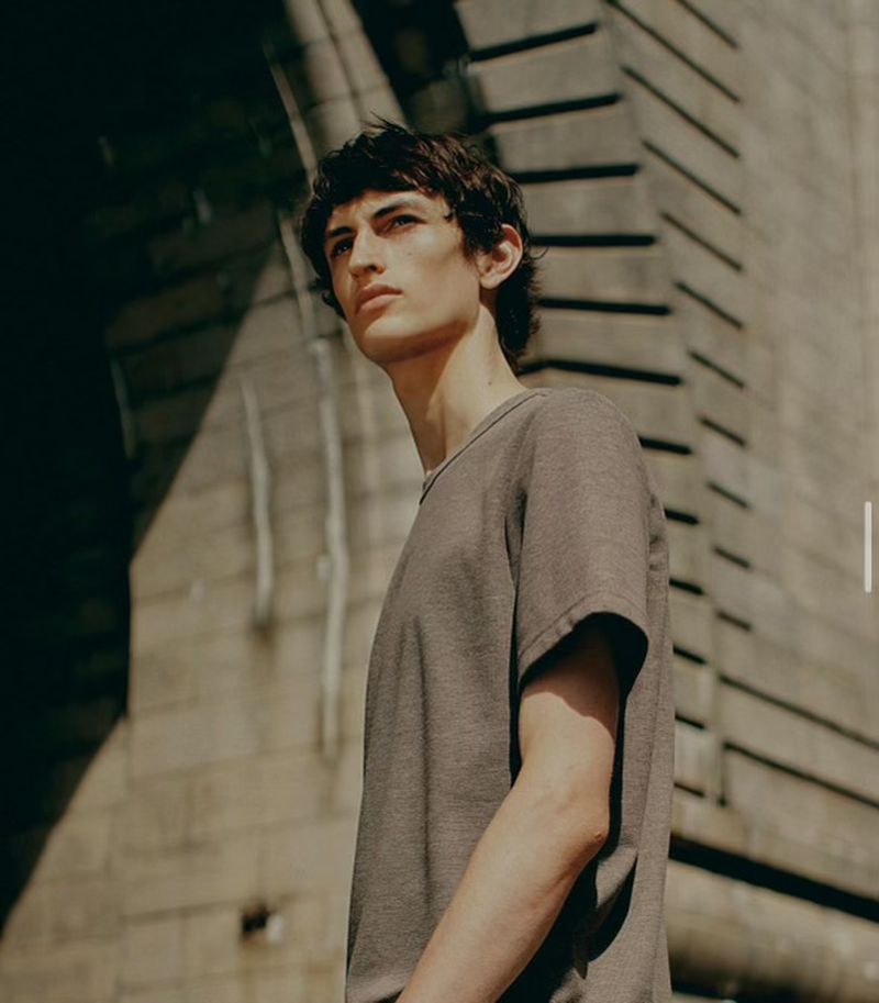 Adult male poses in a short-sleeve shirt in an urban setting under natural light with neutral color grading and stone background.