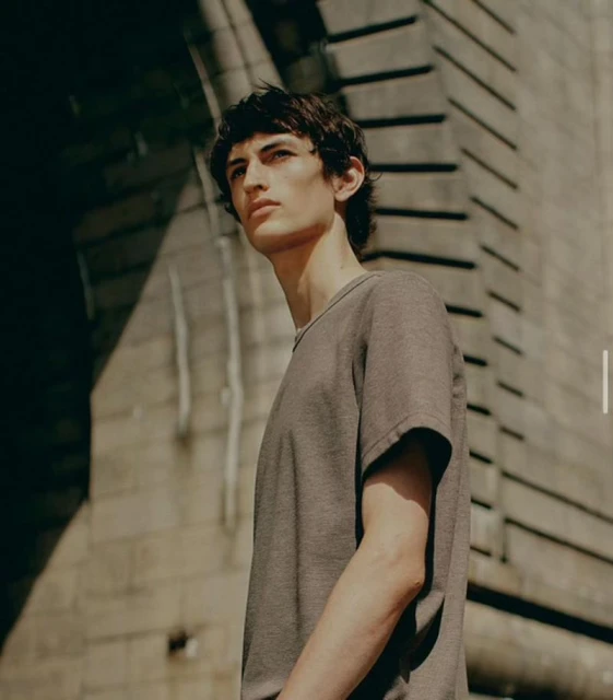 Adult male poses in a short-sleeve shirt in an urban setting under natural light with neutral color grading and stone background.