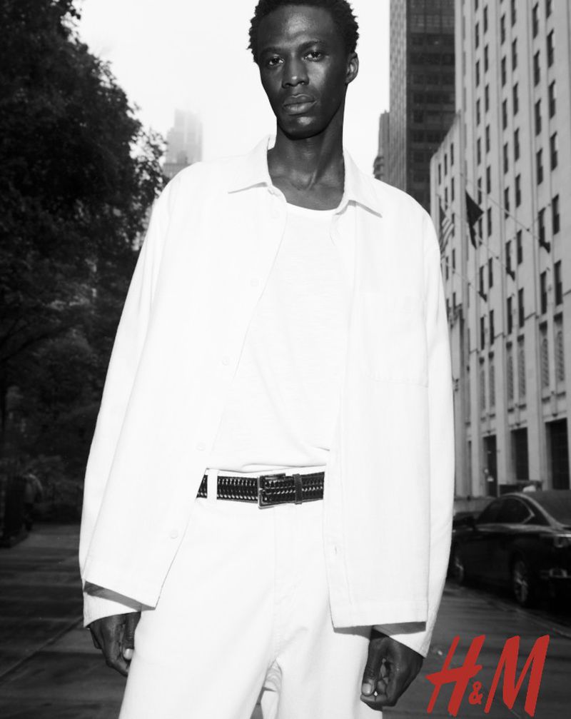 A Black man in an all-white outfit poses outdoors against a city backdrop, with the H&M logo visible at the bottom right.