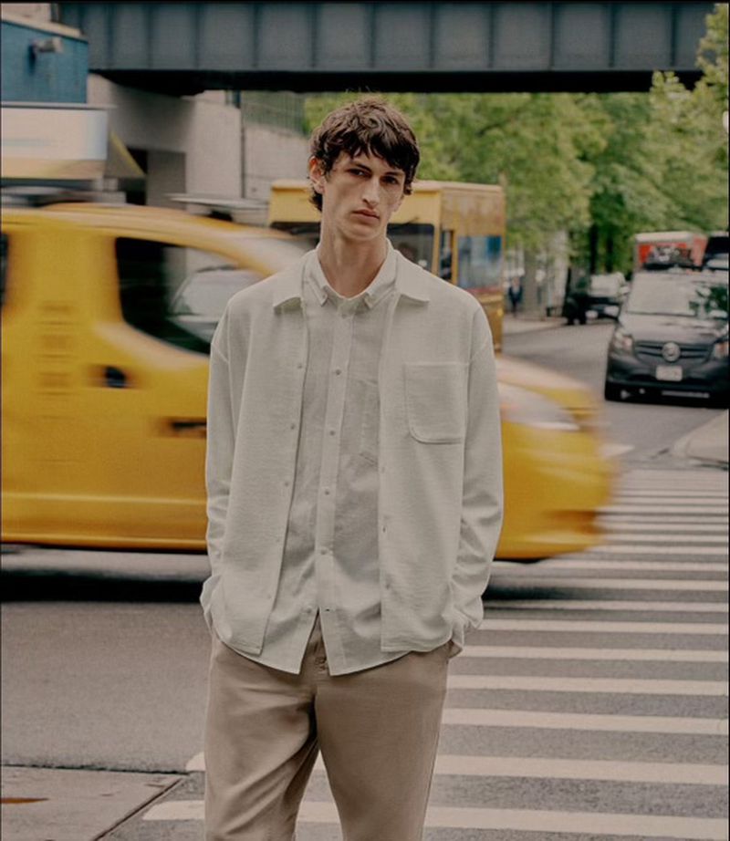Man in white shirt and khaki pants stands on a crosswalk with a yellow taxi blurred in motion in the background in an urban setting.