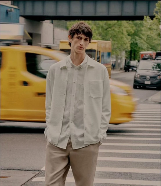 Man in white shirt and khaki pants stands on a crosswalk with a yellow taxi blurred in motion in the background in an urban setting.