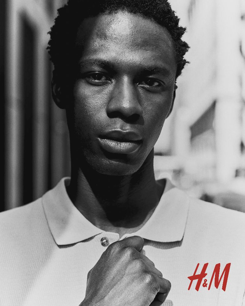 Portrait of a Black male model in a white polo shirt with the H&M logo, photographed outdoors against a blurry building backdrop.