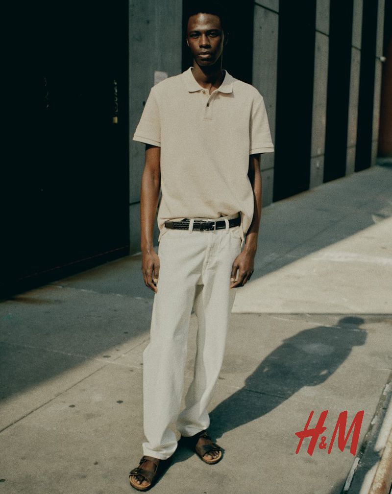 A slender man in a polo shirt and relaxed pants stands on a city sidewalk in the sun, with the H&M logo visible in the bottom corner.