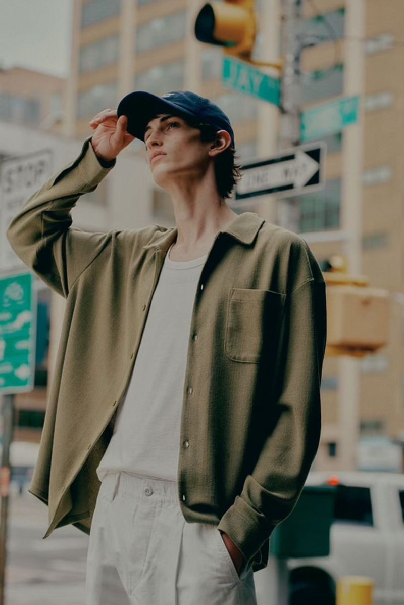 Man in streetwear poses on a city street corner, wearing a baseball cap, olive shirt, white t-shirt, and white pants, illuminated by natural light.