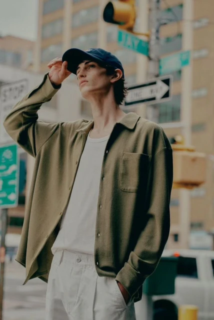 Man in streetwear poses on a city street corner, wearing a baseball cap, olive shirt, white t-shirt, and white pants, illuminated by natural light.