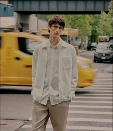 A man in a white button-down shirt and beige trousers standing on a city crosswalk with a blurred yellow taxi behind him.