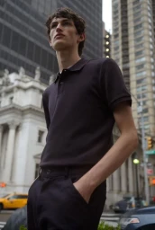 Low angle shot of a male model wearing a dark polo shirt standing outdoors on a city street with tall buildings