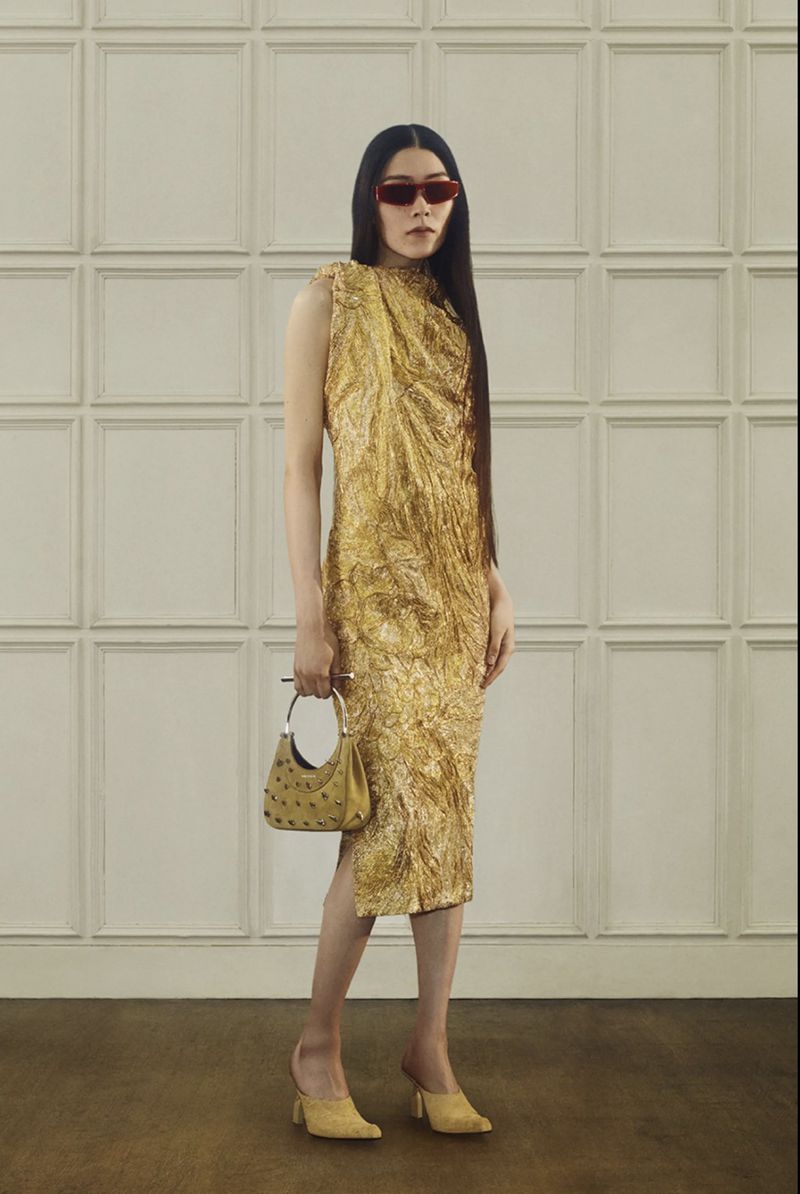 A stylish adult woman in a gold textured midi-dress poses indoors with a structured bag and mules, wearing red sunglasses.