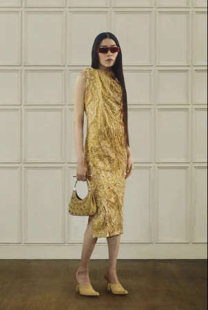 A stylish adult woman in a gold textured midi-dress poses indoors with a structured bag and mules, wearing red sunglasses.