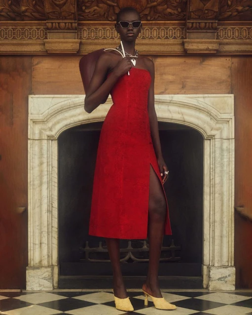 A slender woman with dark skin poses in a red dress and sunglasses, a brown purse over her shoulder, in front of a marble fireplace.