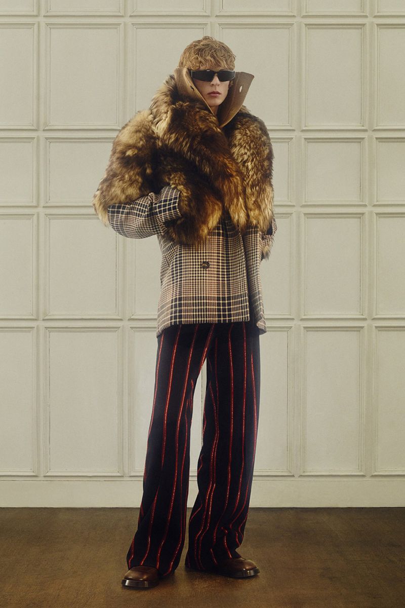 Full shot of person in sunglasses, fur scarf, checkered jacket, and striped pants posing indoors against a paneled wall.