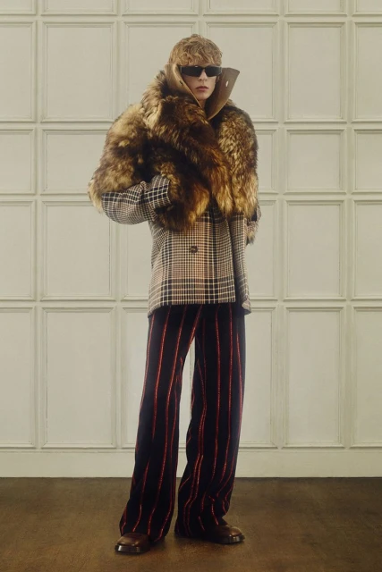 Full shot of person in sunglasses, fur scarf, checkered jacket, and striped pants posing indoors against a paneled wall.