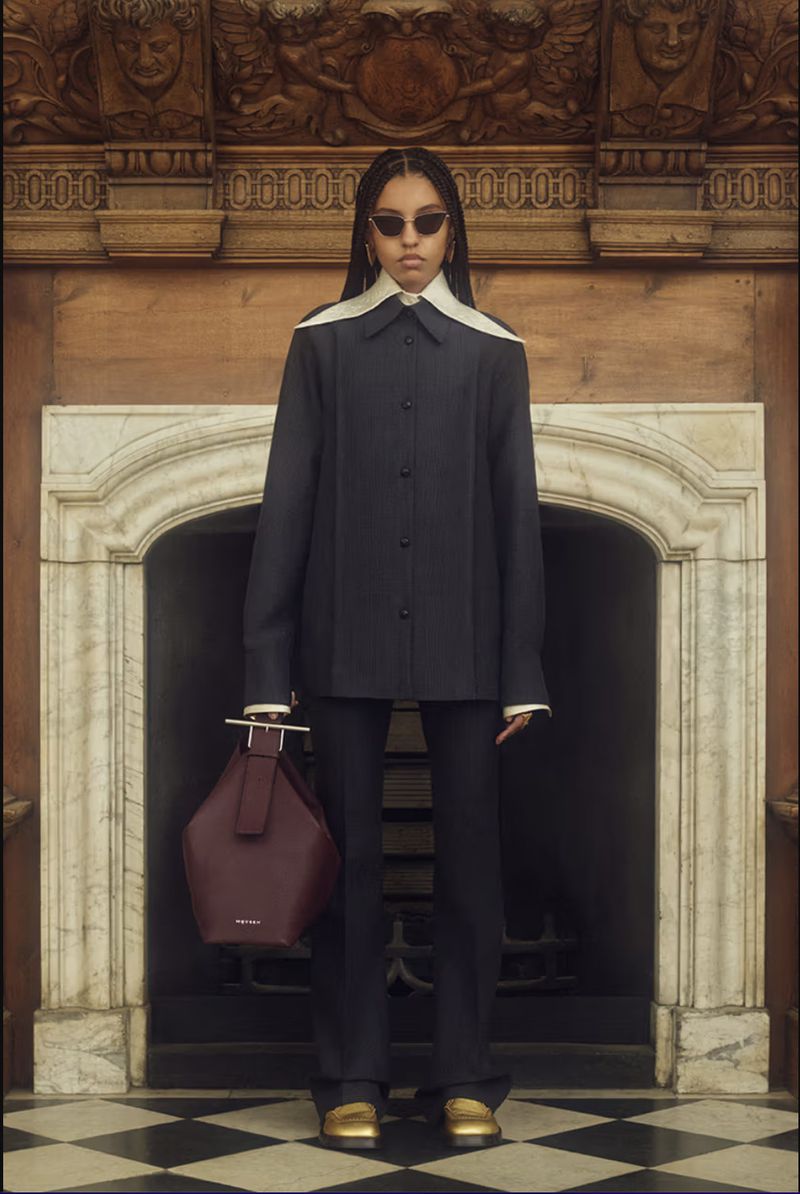 Brunette woman poses in a black tailored suit with an oversized collar, standing in front of a marble fireplace with a maroon handbag.