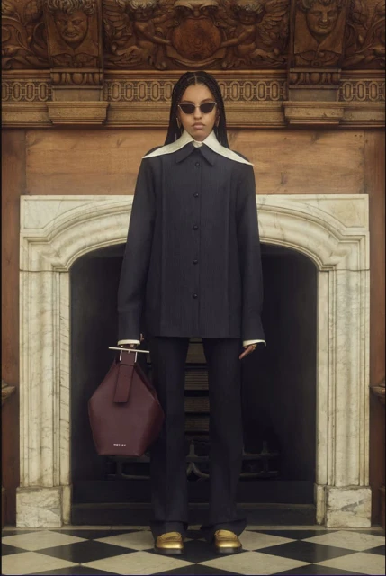 Brunette woman poses in a black tailored suit with an oversized collar, standing in front of a marble fireplace with a maroon handbag.