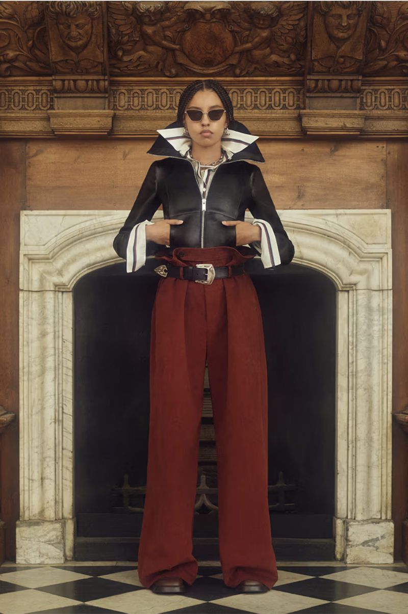 Woman posing indoors wearing a black jacket with a large white collar, red pants, sunglasses, and a belt in front of a fireplace.