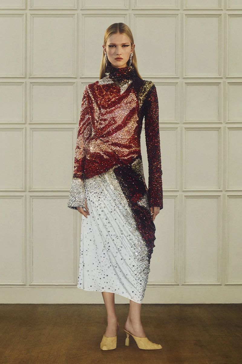 A woman posing in a long-sleeved top with a high neckline, paired with a draped midi skirt and heeled shoes against a panelled backdrop.