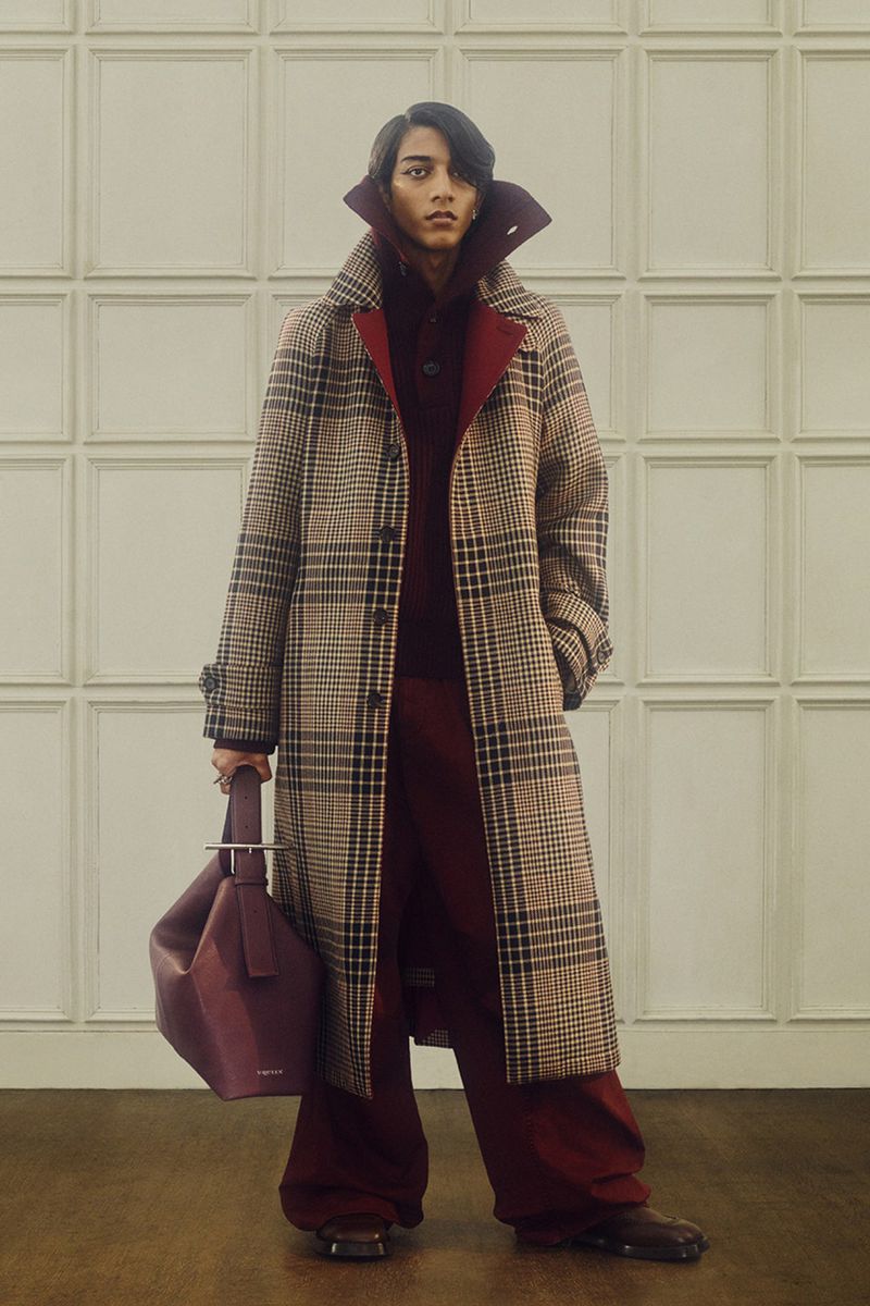 Fashionable individual poses indoors wearing a long plaid coat, maroon sweater and pants while holding a purple bag, looking forward.