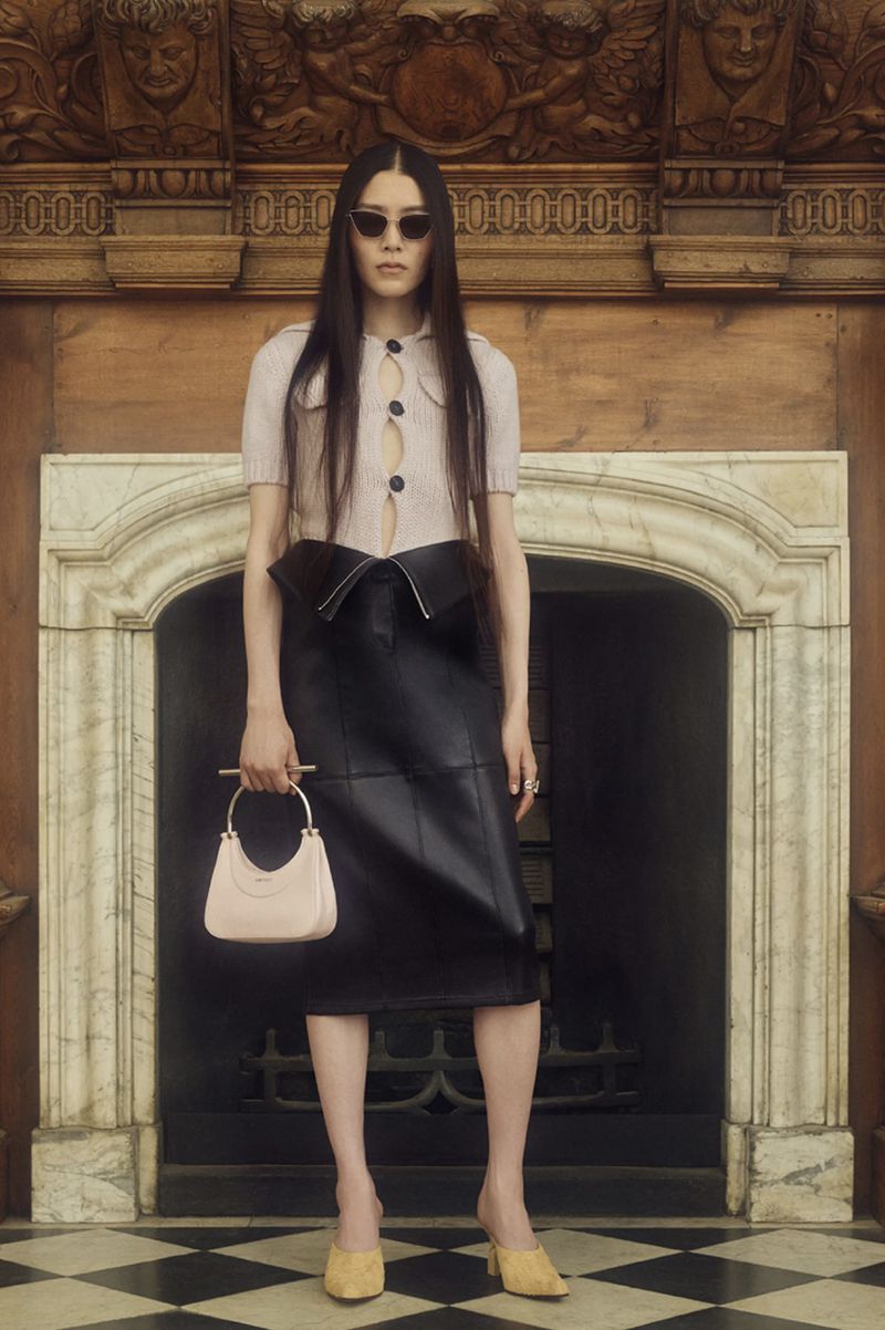 Elegant woman in sunglasses posing in a fashion shot, wearing a knit top, black skirt, handbag, and mules inside a carved-wood interior.