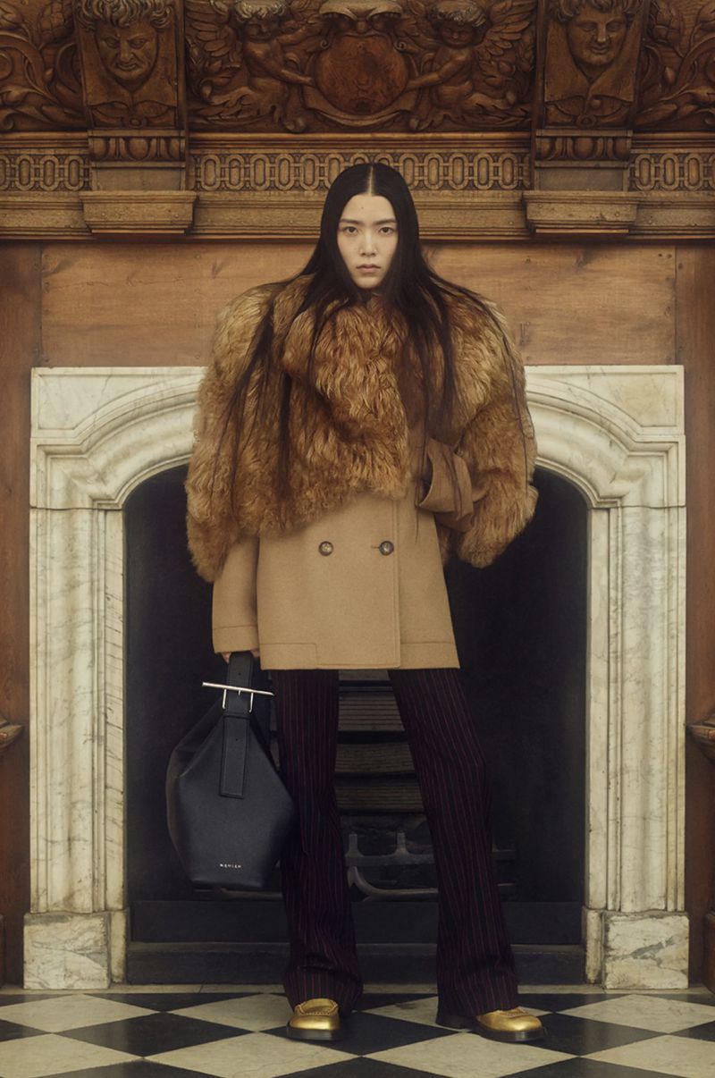 Fashion shot of an Asian woman wearing a fur stole, tan coat, striped pants, and gold shoes, holding a navy bag indoors.