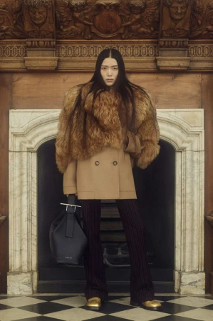 Fashion shot of an Asian woman wearing a fur stole, tan coat, striped pants, and gold shoes, holding a navy bag indoors.