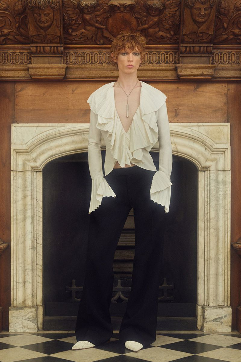 A slender adult model with red hair poses indoors near a decorative fireplace, wearing a ruffled white blouse and black pants.