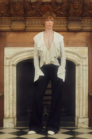 A slender adult model with red hair poses indoors near a decorative fireplace, wearing a ruffled white blouse and black pants.