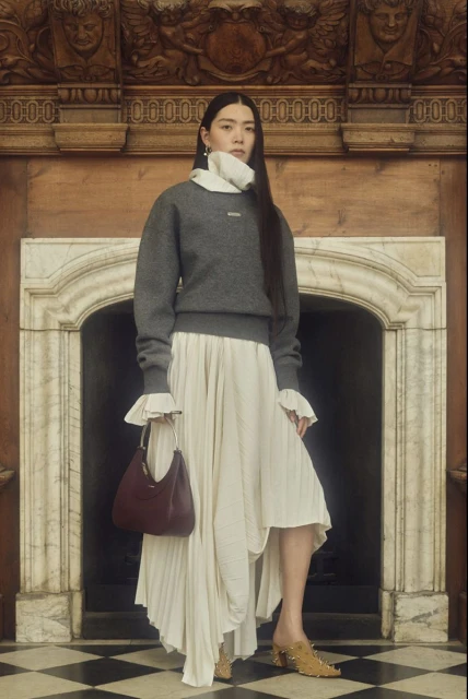 Asian woman in a grey sweater, ivory skirt, and heeled mules, posing in a vintage interior with a small burgundy purse. Fashion editorial.