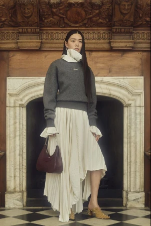 Asian woman in a grey sweater, ivory skirt, and heeled mules, posing in a vintage interior with a small burgundy purse. Fashion editorial.