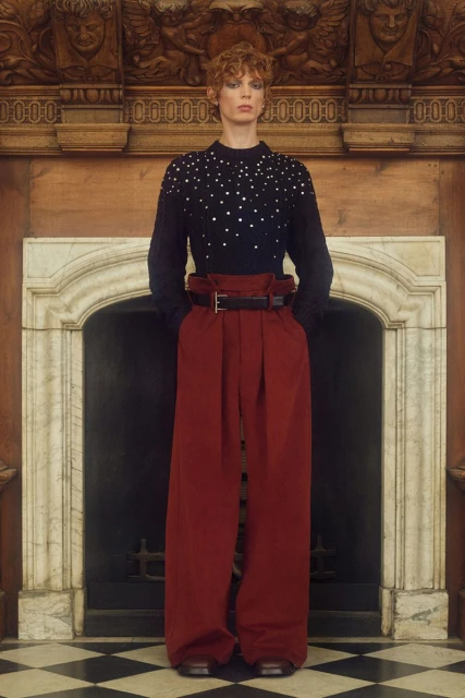 Full-shot fashion portrait of a person with red hair in a black embellished sweater and red wide-leg pants in a marble room.