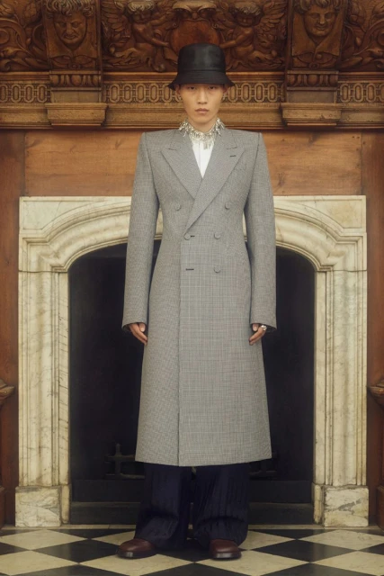 Man in a grey coat and black bucket hat stands in front of a fireplace, creating a striking fashion portrait in an indoor setting.