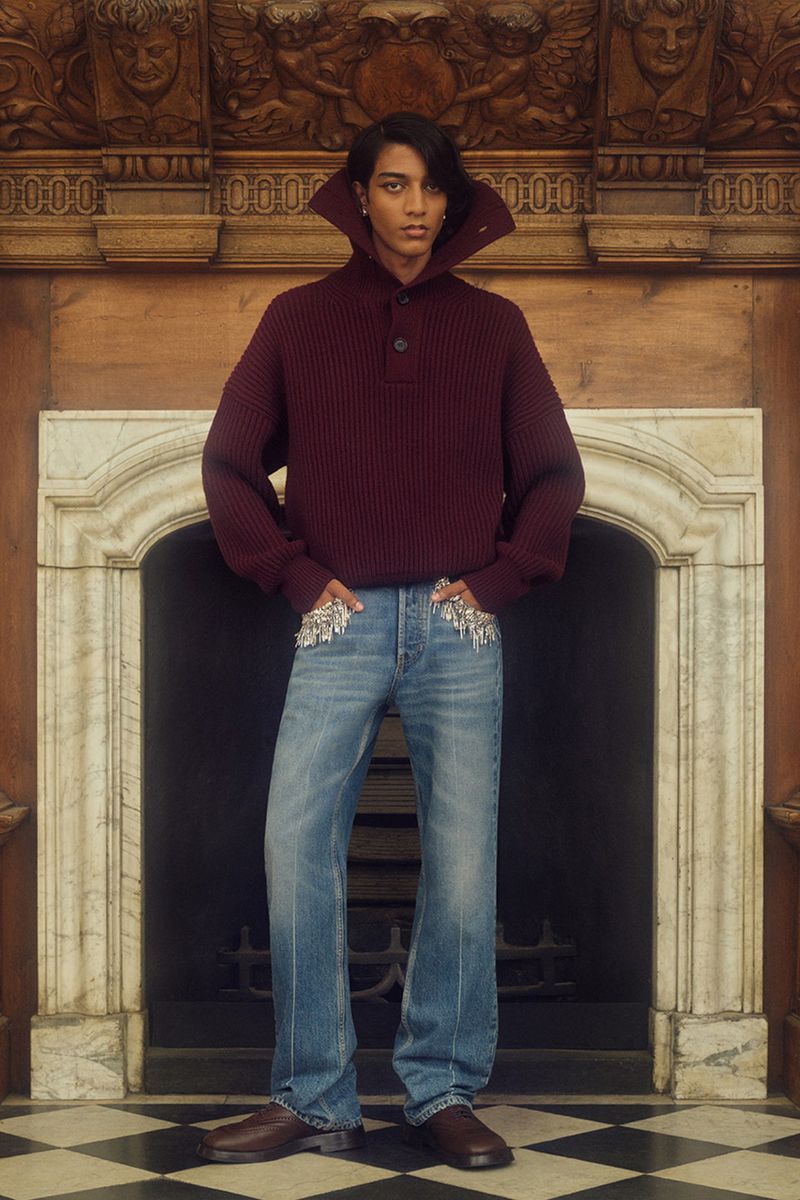 Man poses indoors wearing a maroon sweater and embellished jeans, creating a fashion-forward, elegant, and visually striking image.