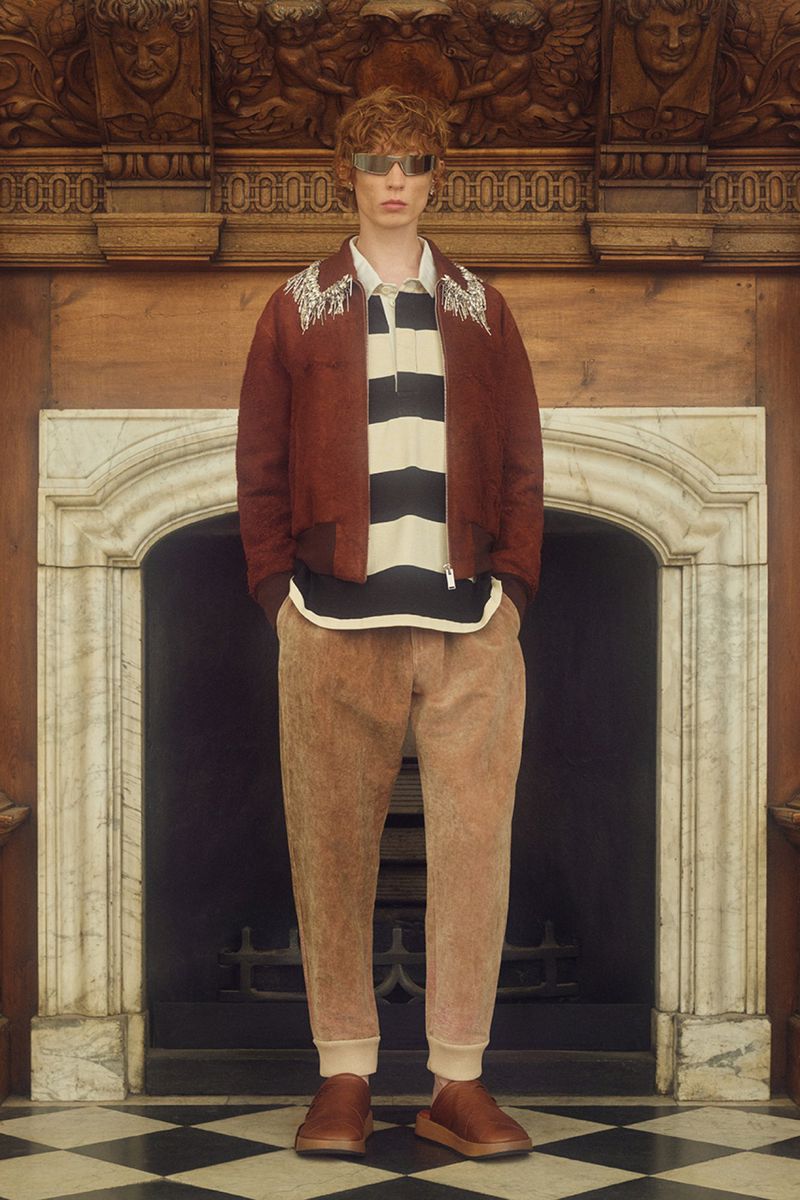A red-haired male model poses in front of a marble fireplace wearing a rust-colored jacket, striped shirt, tan pants, and reflective sunglasses.
