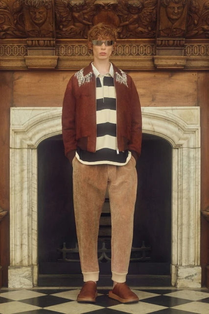 A red-haired male model poses in front of a marble fireplace wearing a rust-colored jacket, striped shirt, tan pants, and reflective sunglasses.