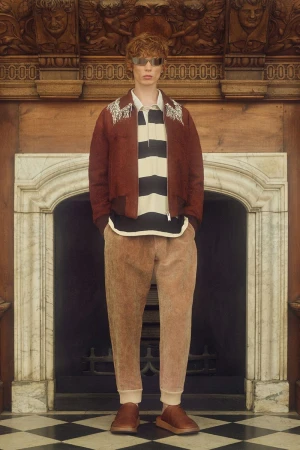 A red-haired male model poses in front of a marble fireplace wearing a rust-colored jacket, striped shirt, tan pants, and reflective sunglasses.