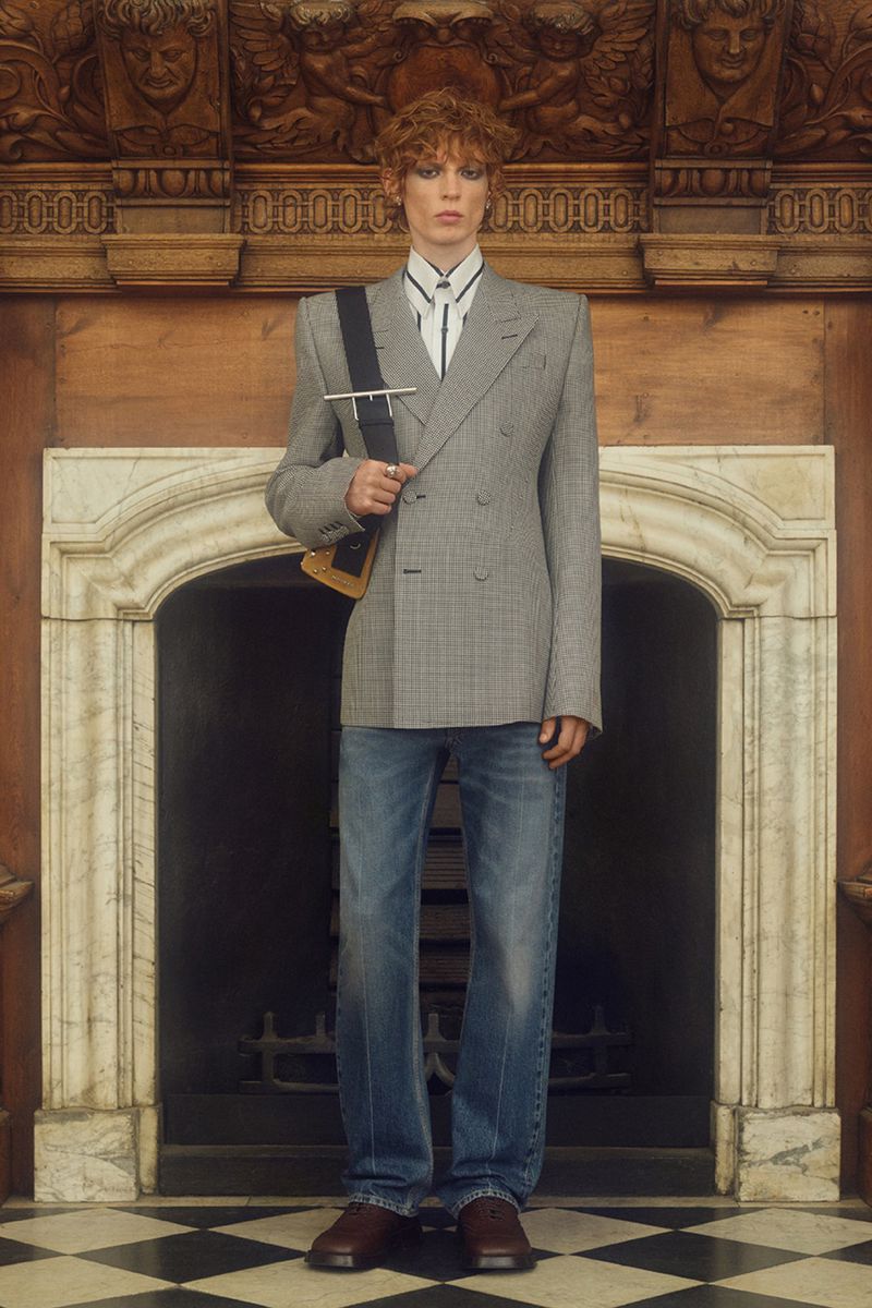 Adult male poses indoors, wearing a grey plaid double-breasted blazer, blue jeans, and brown shoes. Fireplace and wooden details in background.