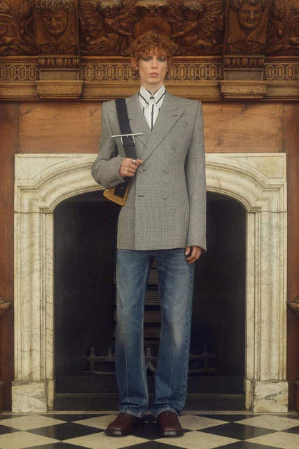 Adult male poses indoors, wearing a grey plaid double-breasted blazer, blue jeans, and brown shoes. Fireplace and wooden details in background.