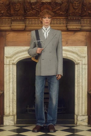 Adult male poses indoors, wearing a grey plaid double-breasted blazer, blue jeans, and brown shoes. Fireplace and wooden details in background.