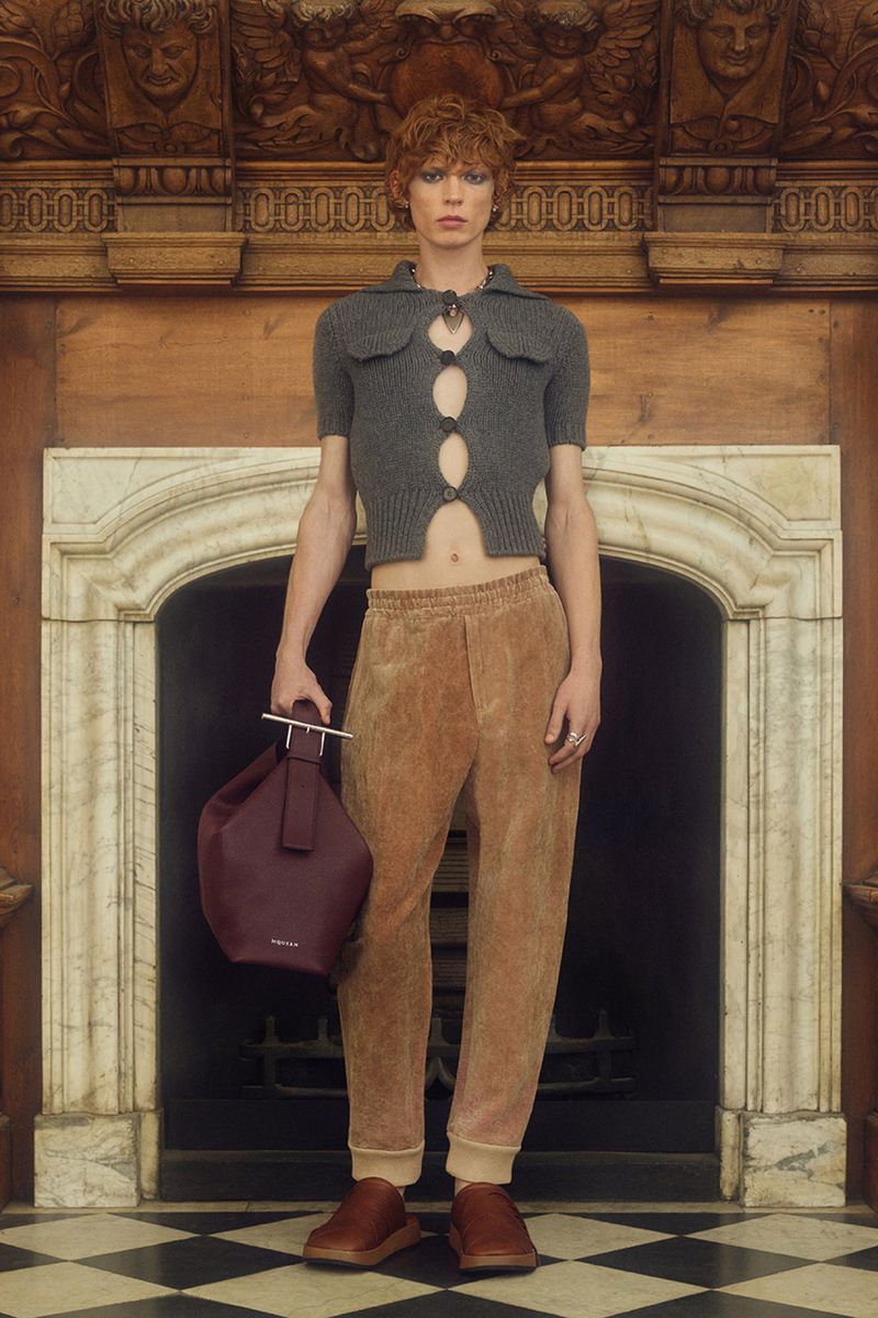 A young adult fashion model in a grey knit top, brown trousers, and brown shoes poses indoors with a burgundy leather bucket bag.