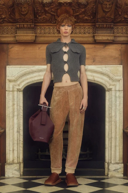 A young adult fashion model in a grey knit top, brown trousers, and brown shoes poses indoors with a burgundy leather bucket bag.