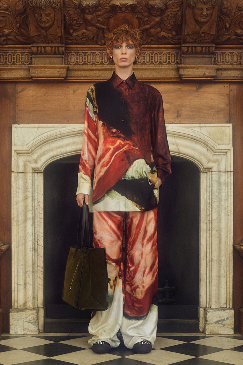 A person in a patterned shirt and pants poses indoors with a tote bag, standing in front of a white marble fireplace and wood details.