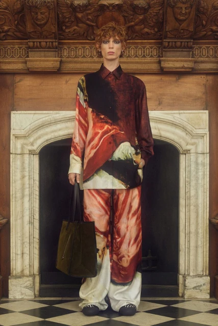 A person in a patterned shirt and pants poses indoors with a tote bag, standing in front of a white marble fireplace and wood details.