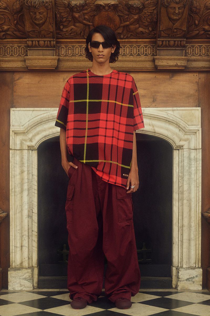 Man in plaid shirt and maroon pants stands in front of an ornate fireplace in an indoor setting with a black and white checkered floor.