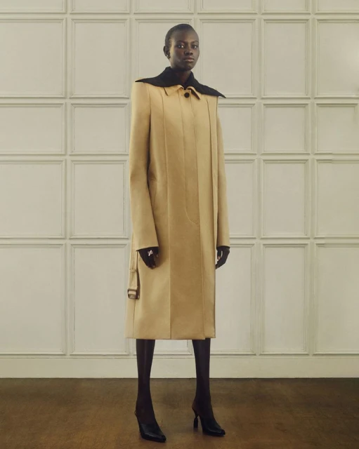 A slender African woman model poses in a tan, calf-length coat with black collar, complementing black tights and heels against a paneled wall.