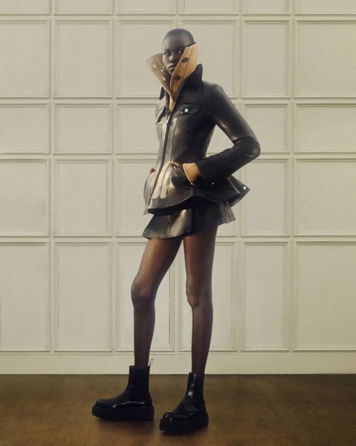 Fashionable young Black woman poses in a stylish black leather jacket and skirt with platform boots in a minimalist indoor setting.
