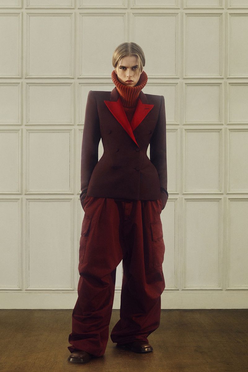 Fashionable adult female model with neutral expression in a burgundy blazer with red trim and oversized red pants, posing indoors.