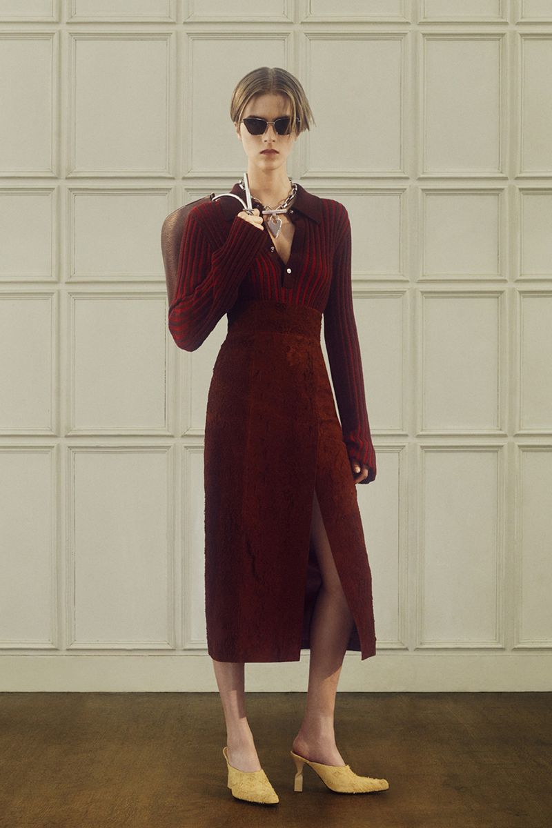 Elegant woman in a burgundy dress, sunglasses, and yellow heels, posing in front of a paneled wall. Shoulder bag, chain necklace.