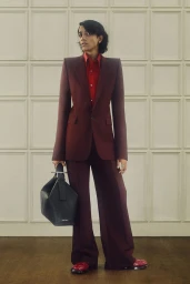Model in a burgundy tailored suit with a red shirt holding a dark handbag against a paneled wall.